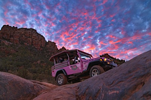 A pink jeep navigates rocky terrain under a vibrant sunset sky with colorful clouds.