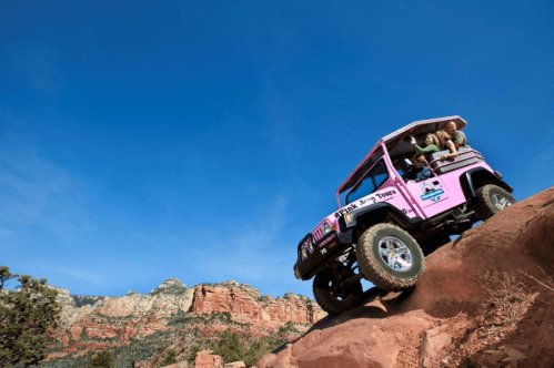 A pink off-road vehicle descends a rocky trail with passengers enjoying the scenic view of red rock formations.