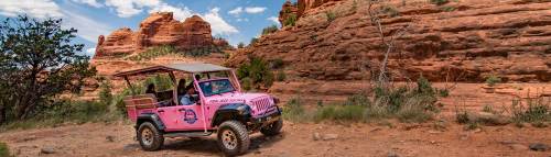 A pink jeep parked on a rocky trail with red rock formations and greenery in the background under a blue sky.