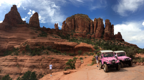 Two pink jeeps parked on red rock terrain with towering rock formations and a blue sky in the background.