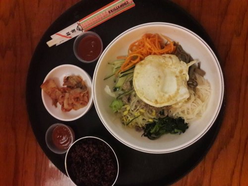 A bowl of bibimbap topped with a fried egg, surrounded by side dishes of kimchi and dipping sauces on a wooden table.