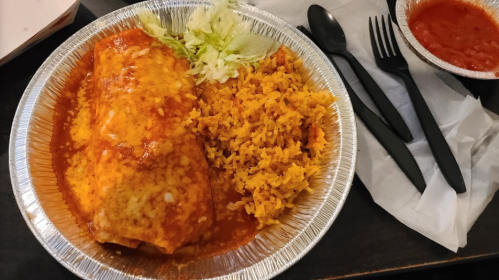 A foil container with a cheesy enchilada, rice, lettuce, and a side of salsa, accompanied by black utensils.