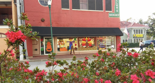 A bakery storefront decorated for fall, with colorful window displays and blooming pink flowers in the foreground.