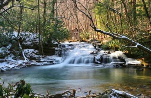 A serene forest scene with a flowing waterfall, surrounded by trees and snow, leading to a small cabin in the background.