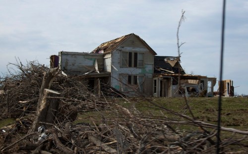 A damaged house with a torn roof, surrounded by fallen trees and debris, under a cloudy sky.