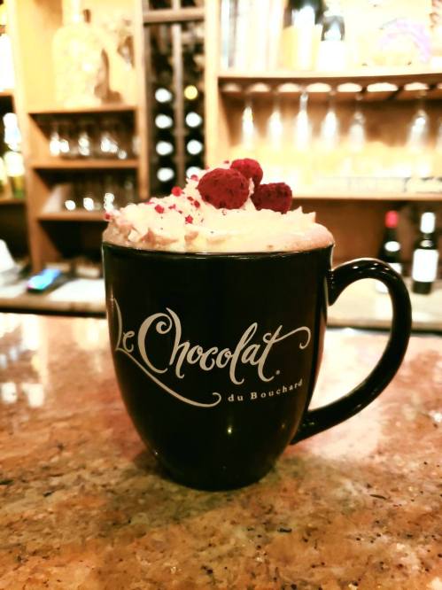 A black mug with "Le Chocolat du Bouchard" logo, filled with whipped cream and topped with red sprinkles and berries.