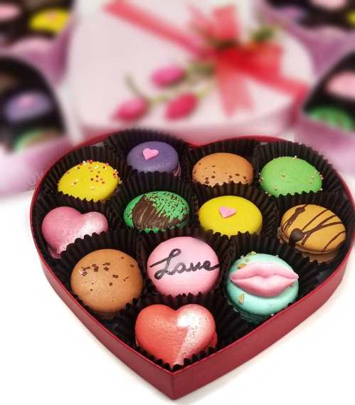 A heart-shaped box filled with colorful macarons, including pink, green, and brown, with decorative designs and a "Love" macaron.