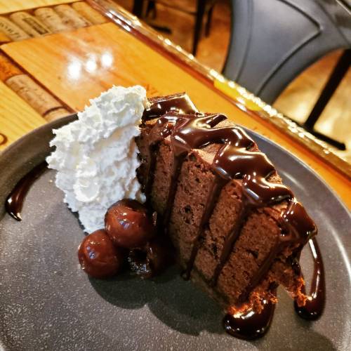 A slice of chocolate cake drizzled with chocolate sauce, served with whipped cream and cherries on a black plate.