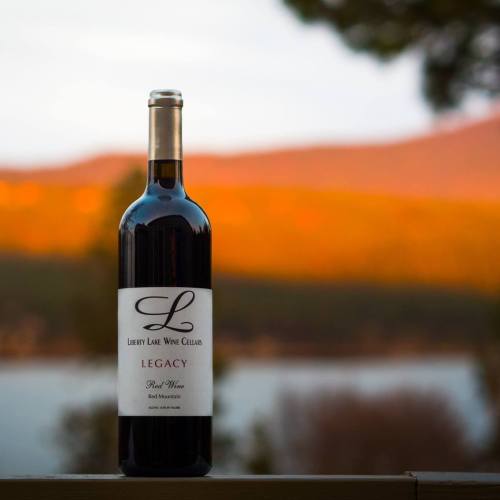 A bottle of Liberty Lake Wine Cellars Legacy red wine, set against a blurred mountain and sunset background.