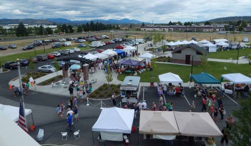 A bustling outdoor market with tents, vendors, and crowds, set against a backdrop of mountains and cloudy skies.