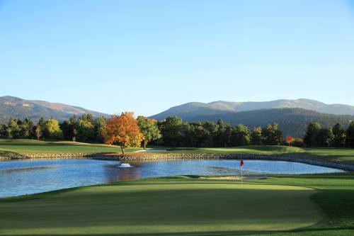 A serene golf course scene with a pond, colorful autumn tree, and mountains in the background under a clear blue sky.