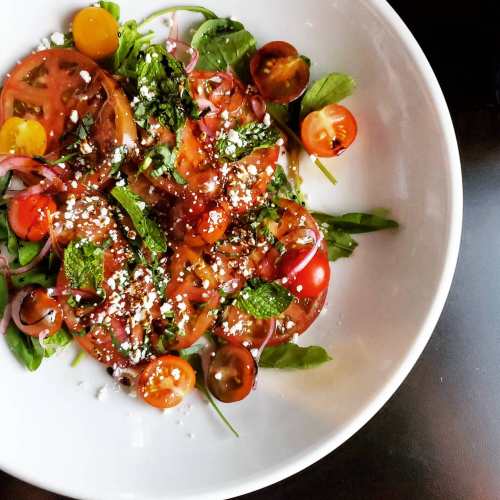 A vibrant salad featuring sliced tomatoes, cherry tomatoes, herbs, and crumbled cheese on a white plate.