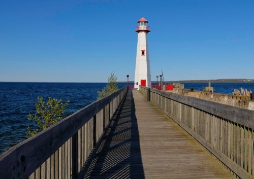A tall white lighthouse with a red roof stands at the end of a wooden pier, overlooking a calm blue sea.