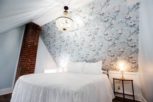 Cozy bedroom with a white bed, floral wallpaper, and a chandelier, featuring a brick accent wall and soft lighting.