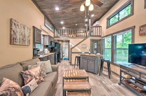 Cozy interior of a modern cabin with a living area, kitchen, and loft, featuring wooden accents and large windows.