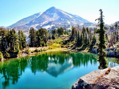A serene mountain landscape with a clear lake reflecting trees and a peak in the background under a bright blue sky.