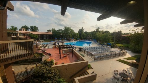 View of a resort pool area with lounge chairs, surrounded by greenery and buildings under a partly cloudy sky.