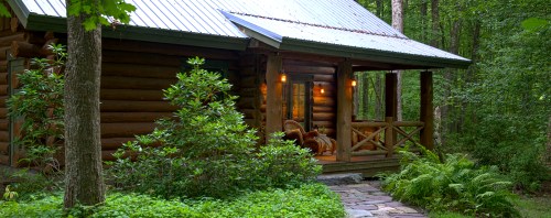 A cozy log cabin surrounded by lush greenery and trees, featuring a welcoming porch and stone pathway.