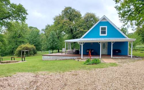 A blue house with a porch surrounded by greenery and gravel, featuring outdoor seating and a fire pit area.