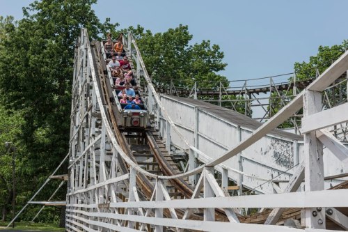 A wooden roller coaster with riders descending a steep drop, surrounded by green trees and a clear blue sky.