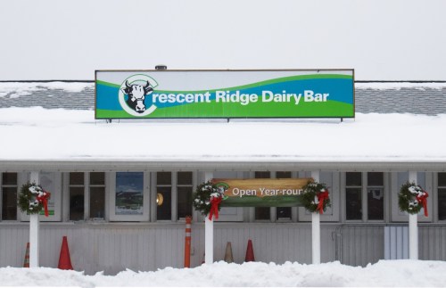Sign for Crescent Ridge Dairy Bar with holiday decorations, snow covering the ground, and a cloudy sky above.