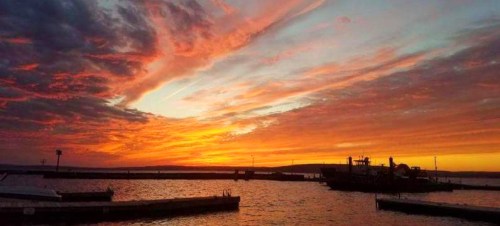 A vibrant sunset over a calm harbor, with colorful clouds reflecting on the water and silhouettes of boats and docks.