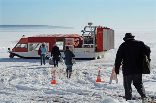 A group of people approaches a red vehicle on a snowy landscape, with orange cones marking the path.