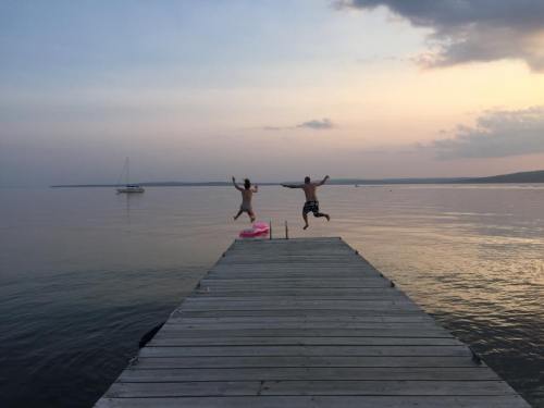 Two people jump off a wooden dock into calm water at sunset, with a sailboat in the background.