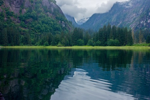 A serene lake surrounded by lush green mountains and trees, with cloudy skies reflecting on the water's surface.