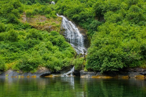 A serene waterfall cascades down a lush, green hillside into a calm, reflective body of water.
