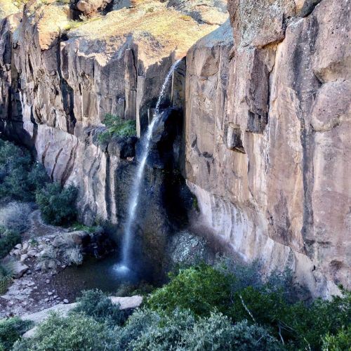 A waterfall cascading down a rocky cliff into a small pool, surrounded by lush greenery and rugged terrain.