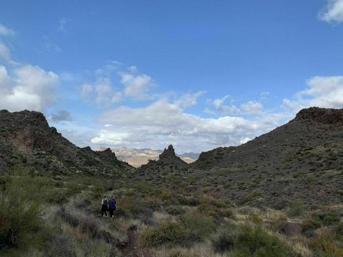 Two hikers walking through a rocky landscape under a blue sky with scattered clouds. Mountains in the distance.