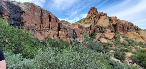 Rocky cliffs rise above lush greenery under a blue sky with wispy clouds.