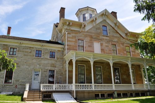 Historic two-story house with a stone exterior, large porch, and a distinctive tower, set against a blue sky.