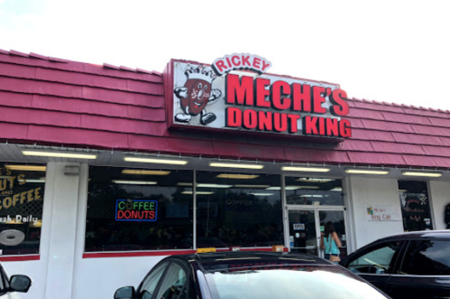 Exterior of Meche's Donut King with a red roof and sign featuring a cartoon donut character. Cars parked outside.