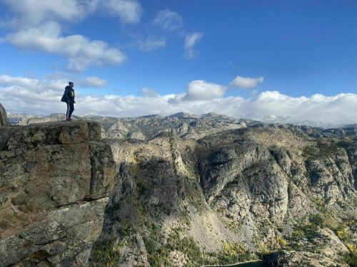 A person stands on a rocky ledge overlooking a vast mountain landscape under a partly cloudy sky.