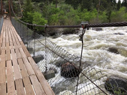 A wooden suspension bridge over a rushing river, surrounded by lush green trees and rocky terrain.