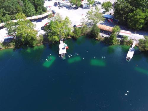 Aerial view of a serene lake with docks, surrounded by trees and parked vehicles along the shore.