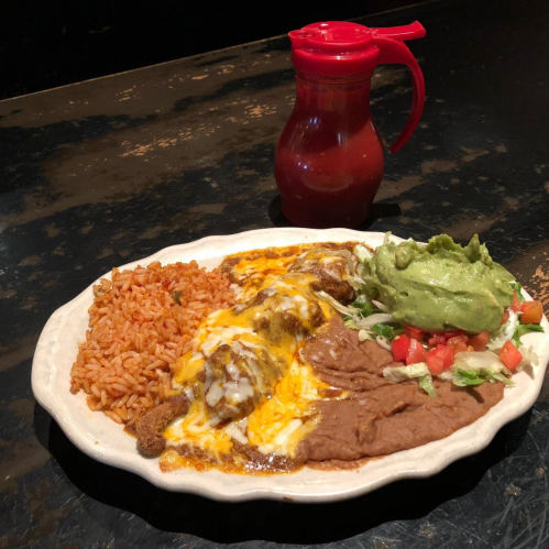 A plate of enchiladas topped with cheese, rice, refried beans, guacamole, and a side of salsa in a red container.