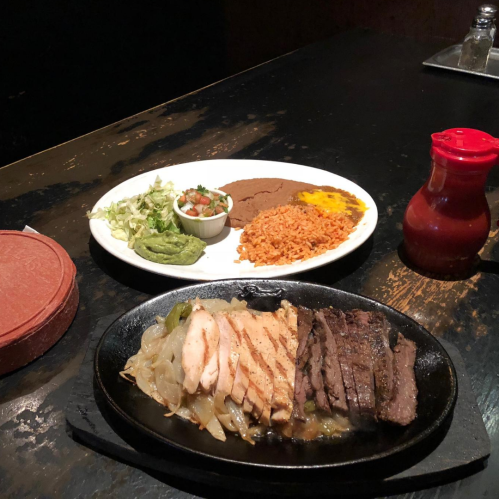 A sizzling platter with grilled chicken and beef, served with rice, beans, salad, and a side of salsa.