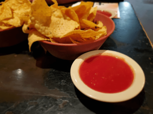 A bowl of tortilla chips next to a small dish of red salsa on a dark table.