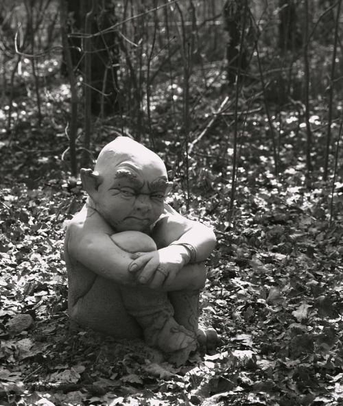 A stone sculpture of a seated figure with exaggerated features, surrounded by a forest of trees and fallen leaves.