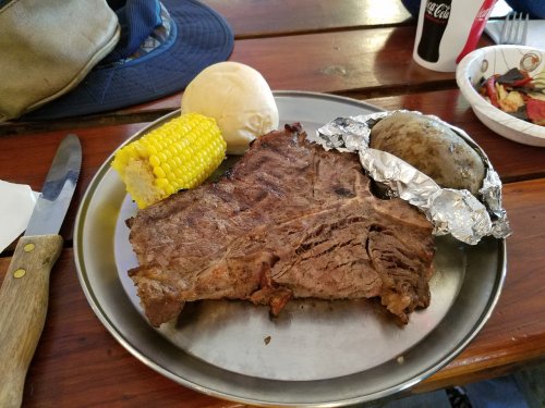 A plate with a large piece of beef, a baked potato in foil, corn on the cob, a roll, and a drink.