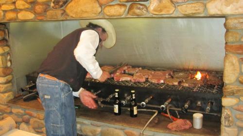 A person in a cowboy hat grills various meats over an open flame, with bottles and a cup nearby.