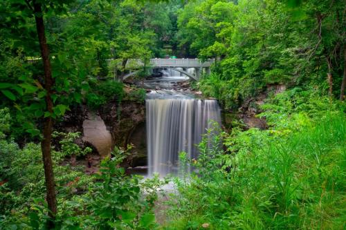 A serene waterfall cascades over rocks, surrounded by lush greenery and a bridge in the background.