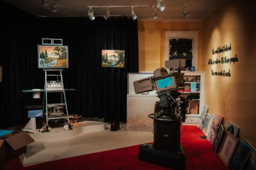 A cozy art studio featuring paintings, a vintage camera, and a wall with text, all set against a dark curtain backdrop.