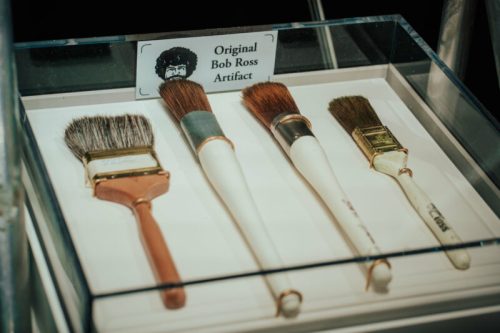 Display of four paintbrushes labeled as original Bob Ross artifacts, showcased in a clear case.