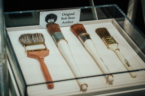 Display of four paintbrushes labeled as original Bob Ross artifacts, with a sign featuring his iconic hairstyle.