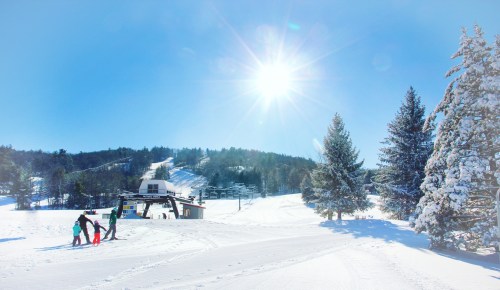 A sunny winter scene with skiers walking towards a ski lift, surrounded by snow-covered trees and slopes.