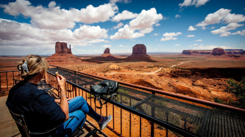 A person sits on a balcony, enjoying a snack while overlooking Monument Valley's iconic rock formations and blue sky.